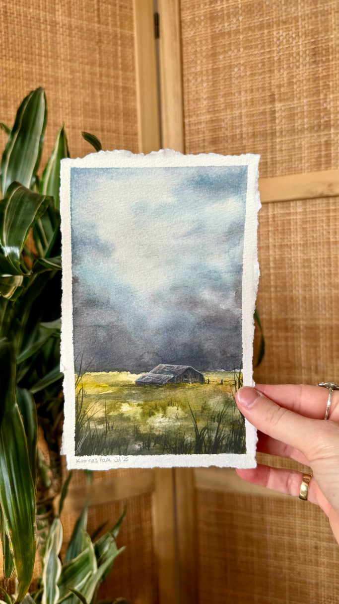 Eine Hand hält eine Aquarellmalerei von einer bewölkten Landschaft mit Wiesen.