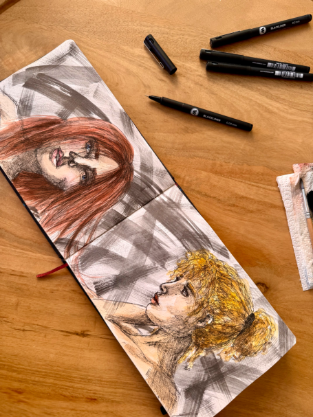 Zwei farbige Porträts von Frauen in einem Skizzenbuch, umgeben von Stiften.