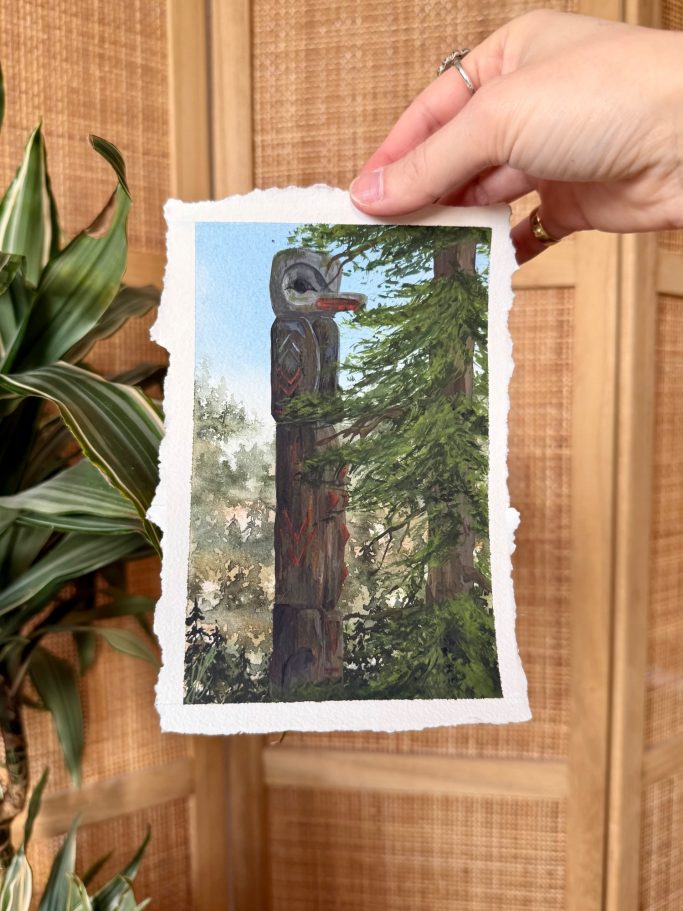 Hand hält ein Foto eines Totempfahls vor Bäumen und Holzverkleidung.