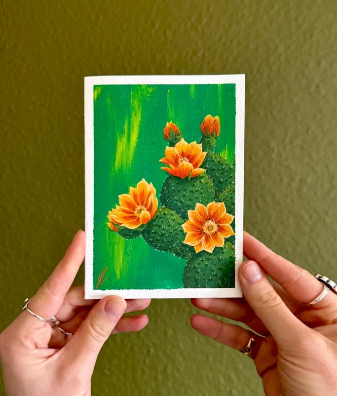 Hände halten eine Postkarte mit einem Kaktus und orangefarbenen Blumen auf grünem Hintergrund.